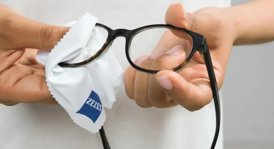 Dłonie czyszczą okulary okularowe białą ściereczką z logo Zeiss.